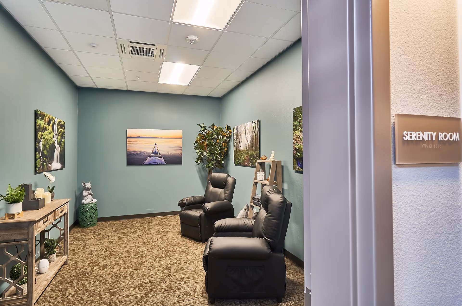 Interior view of a serenity room with two black recliner chairs, a wooden console table with plants and candles, a small green stool with a statue, a ladder shelf with decorative items, and nature-themed wall art on teal walls.