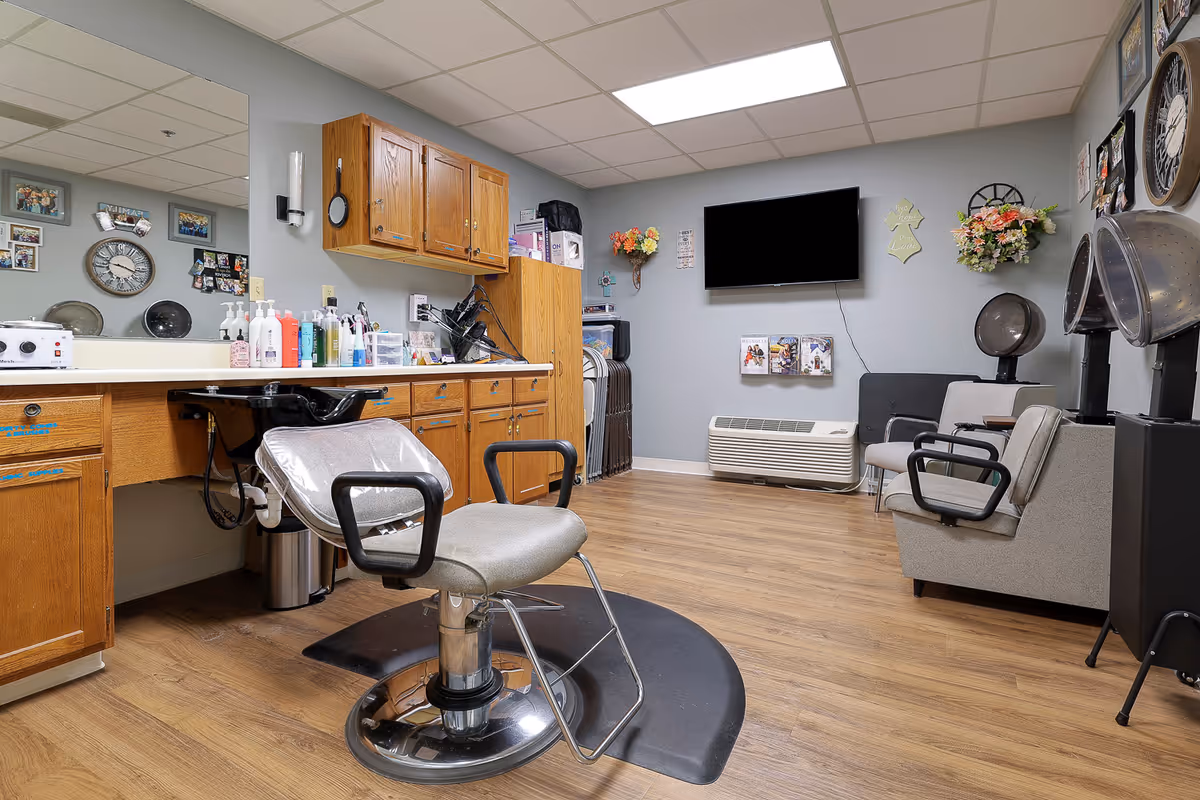 Interior view of a hair salon area in an assisted living facility featuring a salon chair in front of a sink and wooden cabinets. The room has a wooden floor, a wall-mounted TV, hair drying chairs, and decorative wall hangings including flowers and clocks.