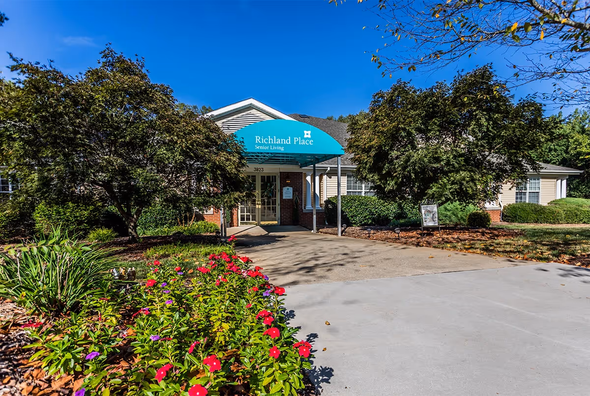 Front exterior view of a senior living facility named Richland Place with a blue awning over the entrance. The building is surrounded by green trees and colorful flowers along the walkway leading to the entrance.