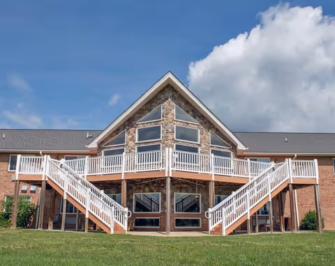 Brick senior living building exterior featuring a central stone A-frame with large triangular windows and symmetrical double staircases up to a white raised deck.