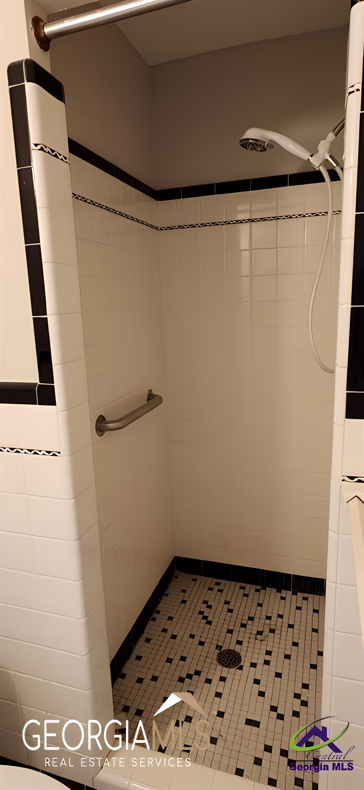 A tiled walk-in shower with white and black tiles. The shower has a handheld showerhead attached to the wall and a metal grab bar for support. The floor is tiled with small square tiles in a black and white pattern.