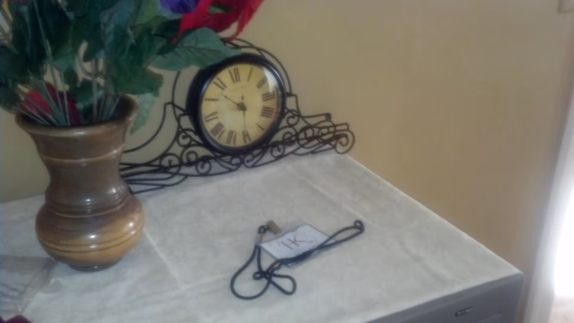A decorative table with a brown vase holding artificial flowers, a vintage-style clock with Roman numerals on a black ornate metal stand, and a lanyard with a badge labeled '1K' lying on the surface.