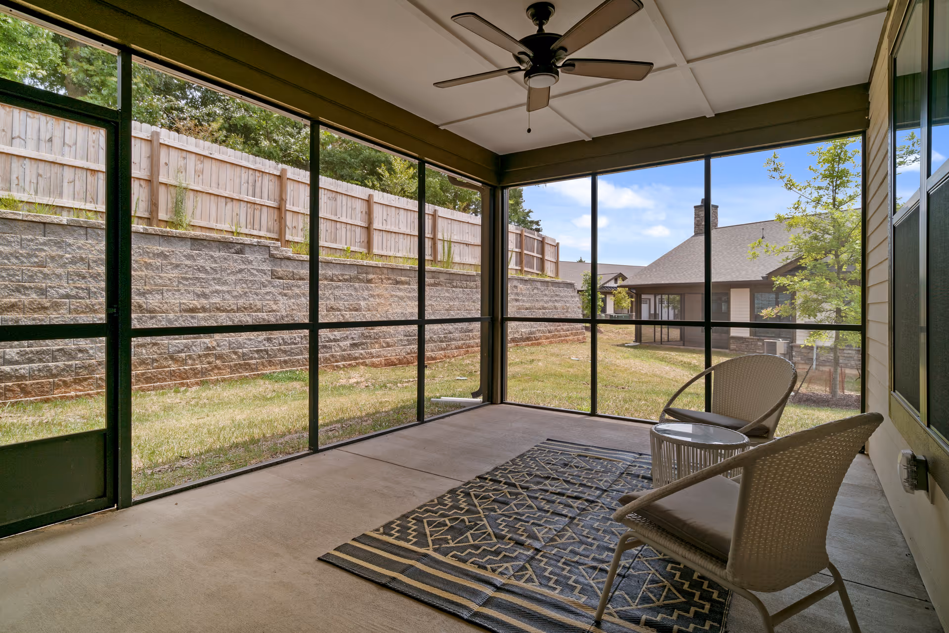 A screened-in porch with two wicker chairs and a small round table on a patterned rug. The porch overlooks a grassy yard with a retaining wall and wooden fence, and neighboring buildings are visible in the background under a partly cloudy sky.