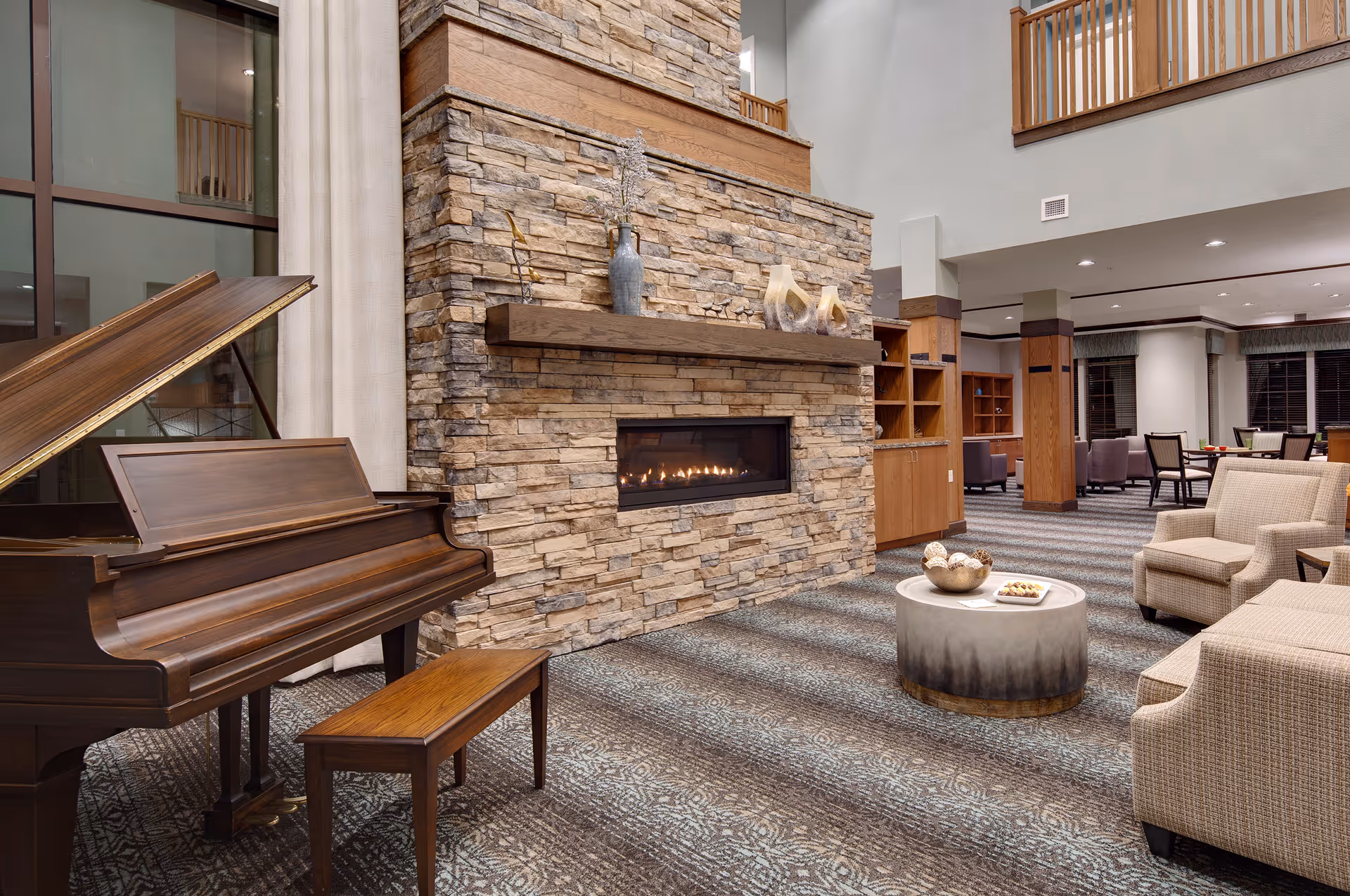 A cozy living room area in a senior living facility featuring a grand piano with a bench, a stone fireplace with a wooden mantel decorated with vases and sculptures, and comfortable upholstered chairs around a round coffee table with decorative items. The room has a patterned carpet and wooden accents, with an open second-floor balcony visible above.