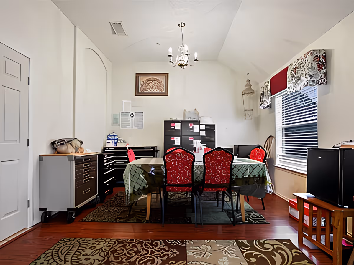 Interior view of a room with a dining table covered with a green patterned tablecloth and surrounded by red cushioned chairs. The room has wooden flooring with multiple area rugs, a window with blinds and a decorative valance, filing cabinets, and a chandelier hanging from the ceiling. There are various items on the cabinets and walls, including a framed picture and some papers.
