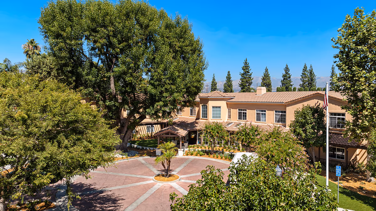 Front exterior view of Ivy Park at West Hills senior living facility with a circular driveway, surrounded by large trees and landscaping under a clear blue sky.