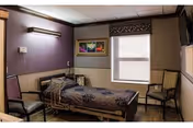 A small, neatly arranged patient room in a skilled nursing facility featuring a hospital bed with patterned bedding, two chairs, a window with a decorative valance, a wall-mounted television, and framed artwork on the wall.