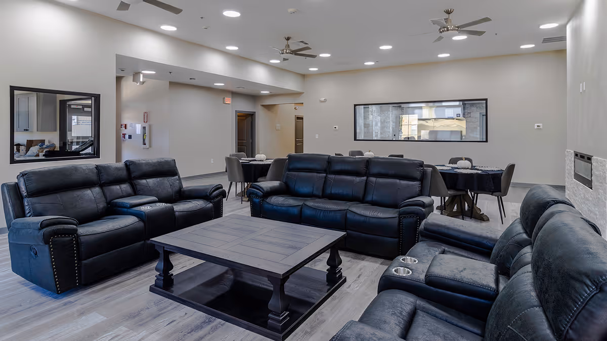 A spacious living room area with three black leather sofas arranged around a large wooden coffee table. The room has light-colored walls, a ceiling fan, recessed lighting, and a large window on one wall. In the background, there are dining tables with chairs and a hallway leading to other rooms.