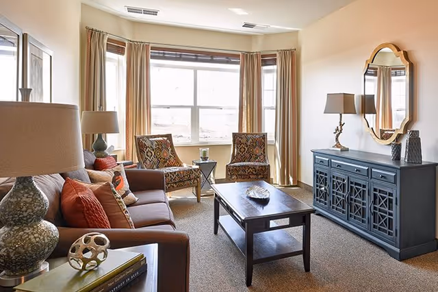 Sunlit living room with a sofa, patterned armchairs by a large bay window, a coffee table, and a blue console topped by a mirror.