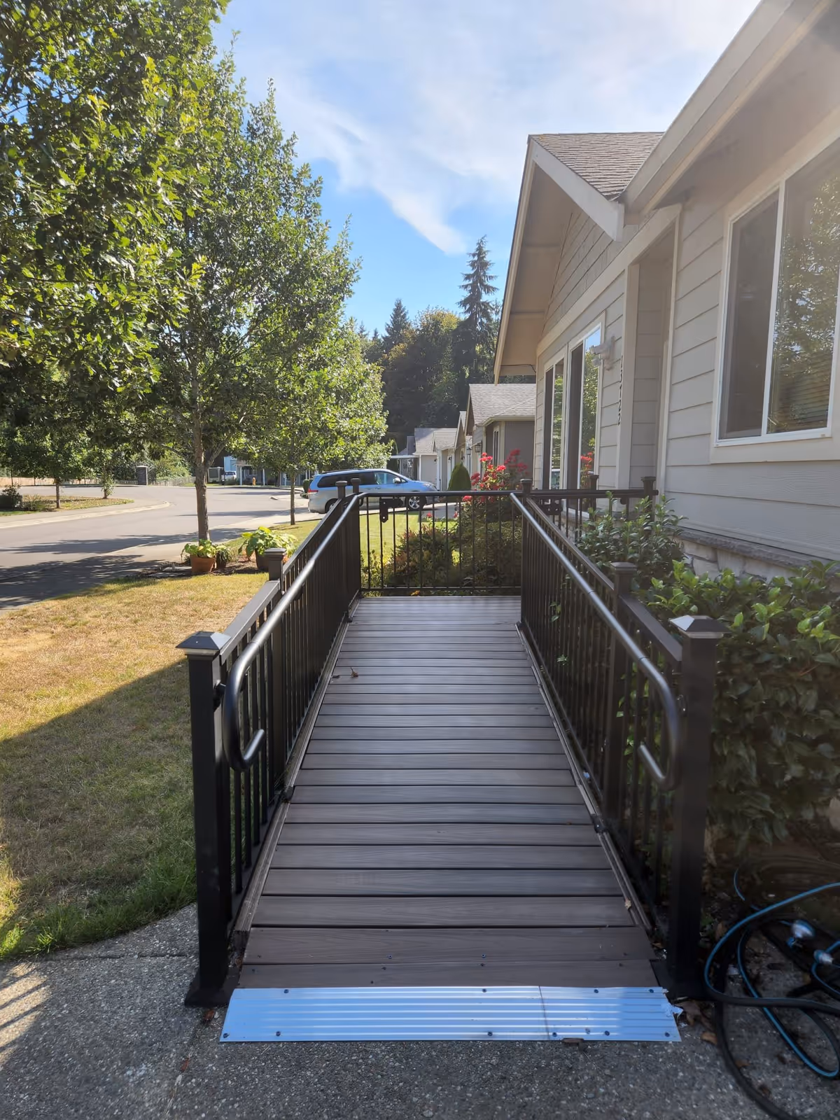 A wooden wheelchair ramp with black metal railings leading to the entrance of a single-story building. The building has light gray siding and several windows. There are green bushes and red flowers near the entrance, a tree on the left side, and a parked silver car in the background under a blue sky with some clouds.