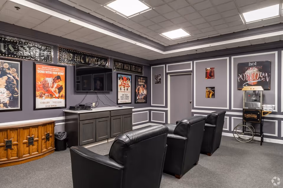 A cozy entertainment room with three black leather armchairs facing a wall-mounted flat-screen TV. The walls are decorated with vintage movie posters and signs, including one that says 'hot fresh popcorn' next to a popcorn machine on wheels. The room has gray walls with white trim and a gray carpeted floor.