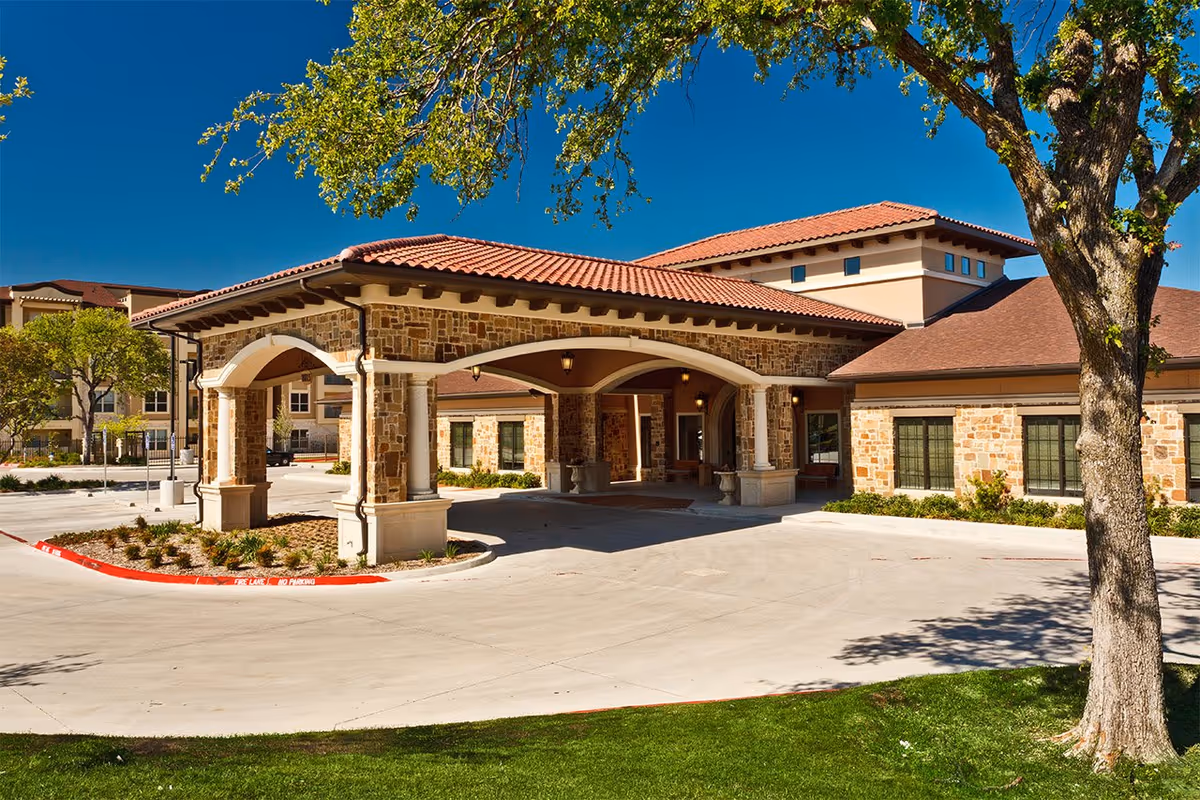 Exterior view of The Villages of Lake Highlands senior living facility showing a stone and stucco building with a covered entrance, red tile roof, and surrounding trees and landscaping under a clear blue sky.