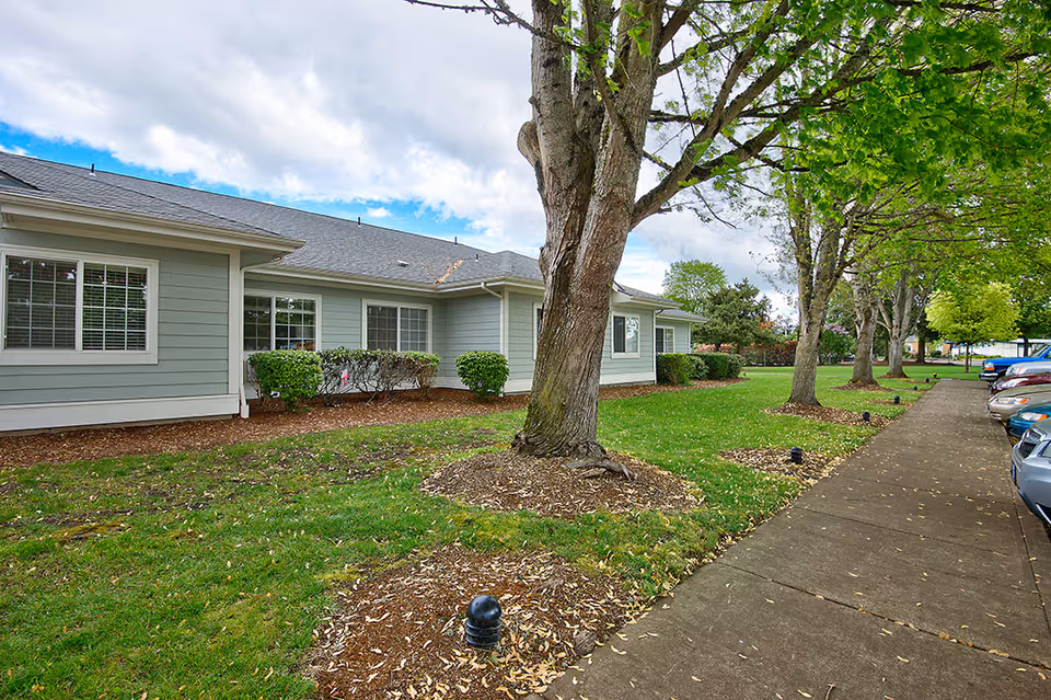 Exterior view of a single-story assisted living facility building with light gray siding and white trim. The building is surrounded by green grass, trees, and shrubs. A sidewalk runs alongside the building with parked cars visible on the right side. The sky is partly cloudy.
