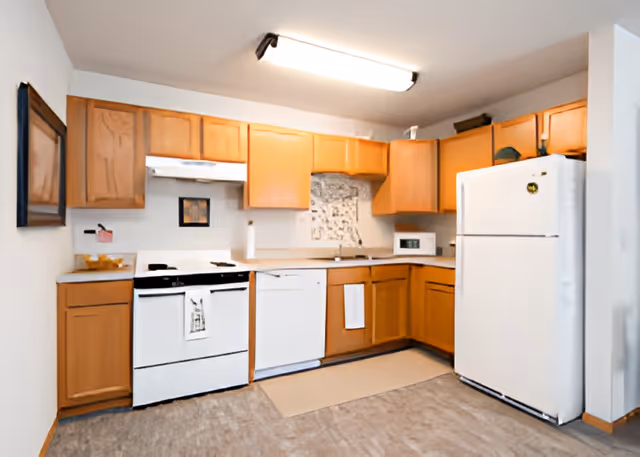 A kitchen with wooden cabinets, a white refrigerator, a white stove with an oven, a white dishwasher, a microwave, and a countertop with a sink. The floor is covered with a light-colored mat in front of the sink area, and there is a fluorescent ceiling light fixture.