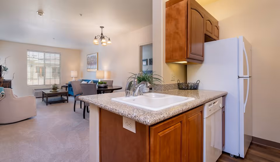Open-plan apartment interior with a kitchen island and sink in the foreground and a connected living/dining area with sofa and table in the background.