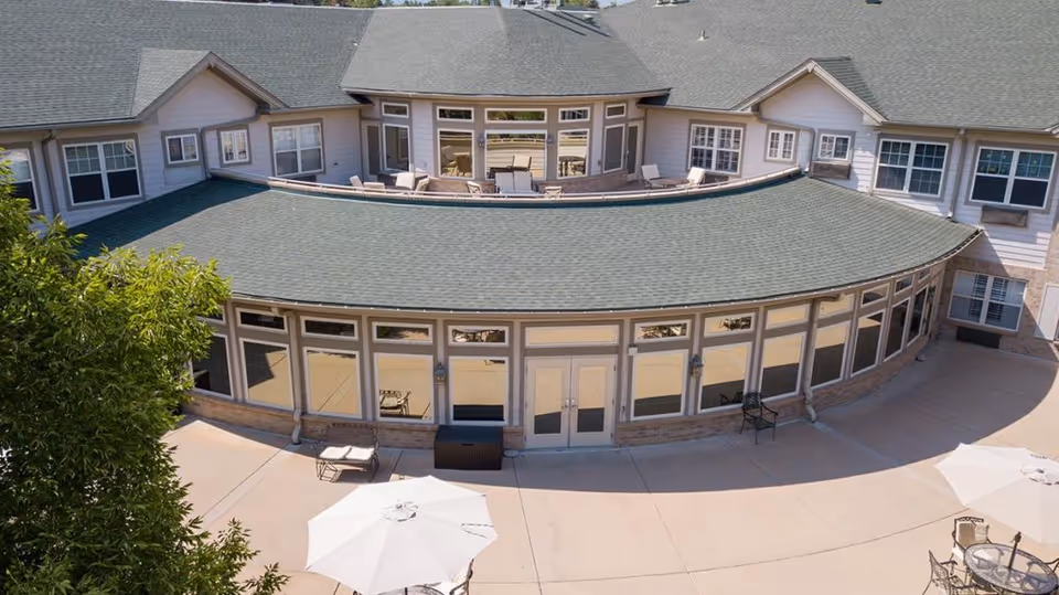 Curved two-story senior living building with large windows overlooking a paved courtyard with umbrellas and patio furniture.