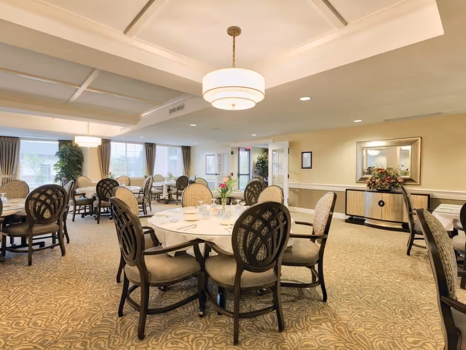 A well-lit dining room with round tables covered with white tablecloths, each surrounded by cushioned chairs with dark wooden frames. The room has beige walls, patterned carpet, large windows with curtains, and decorative plants. A sideboard with a floral arrangement and a large mirror is visible on one wall.