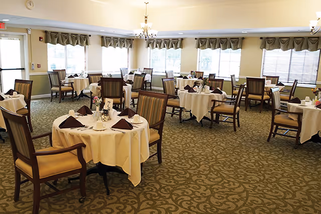 A spacious dining room with multiple round tables covered with white tablecloths, each set with folded brown napkins, plates, and small flower arrangements. The room has large windows with valance curtains, patterned carpet, and wooden chairs with cushioned seats and backs. Ceiling lights and chandeliers provide illumination.