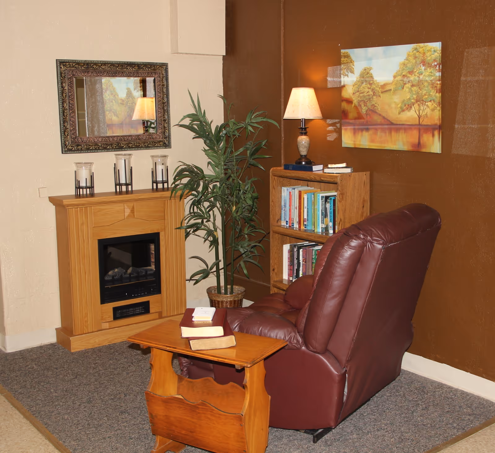 Cozy reading nook featuring a maroon recliner, wooden side table, bookshelf, lamp, potted plant and faux fireplace.