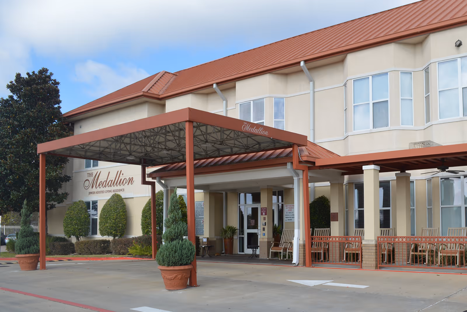 Exterior view of The Medallion Jewish Assisted Living facility showing the entrance with a covered drop-off area, potted plants, and a seating area with chairs on the porch. The building has beige walls, large windows, and a red roof.
