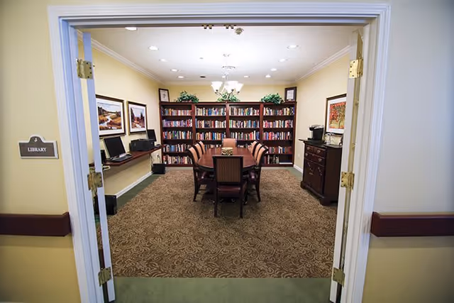 View through double doors into a library room with a rectangular table surrounded by eight chairs. The back wall has bookshelves filled with books and decorative plants on top. On the left wall, there are two framed pictures above a desk with two computers. On the right side, there is a wooden cabinet with a coffee maker and a framed picture above it. The room has beige walls, a patterned carpet, and ceiling lights.