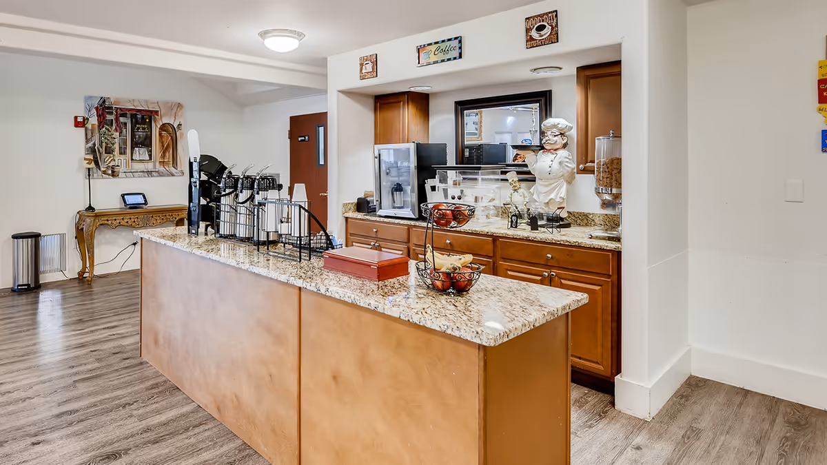 Granite-topped coffee and refreshment bar with beverage dispensers, cabinets, and decor in a senior living facility common area.