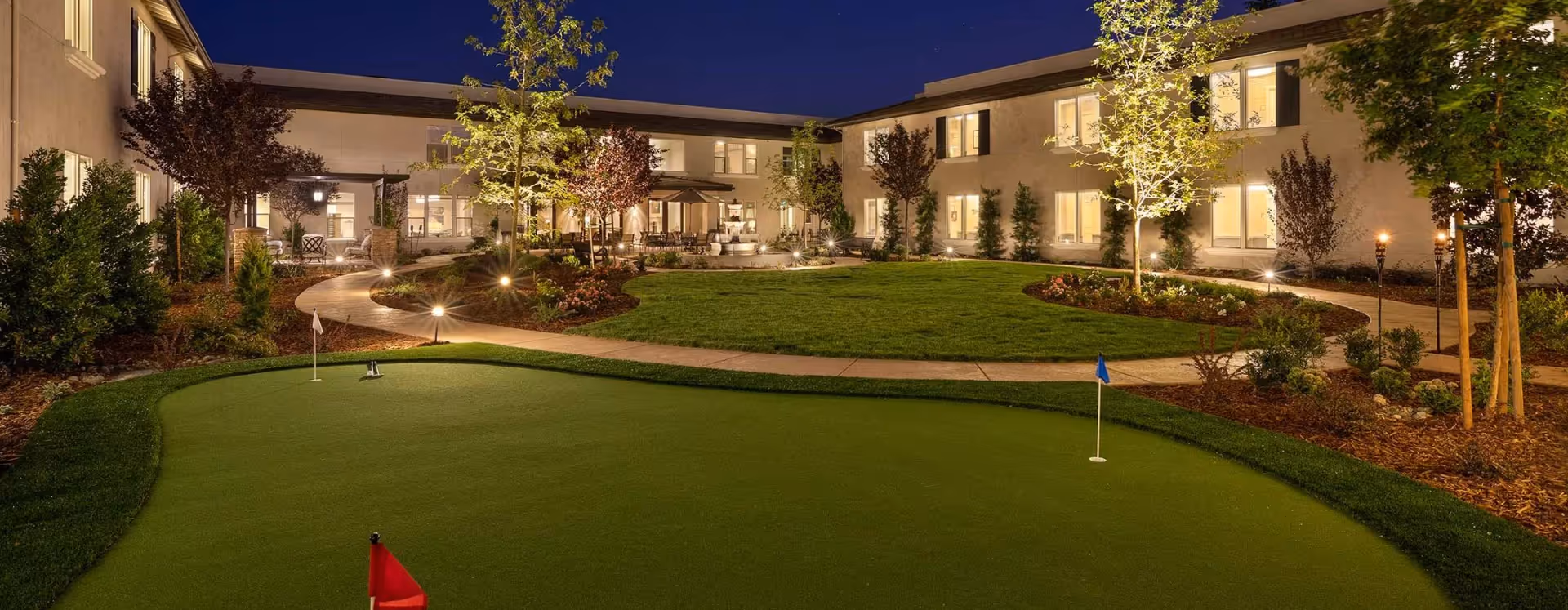 Night view of a landscaped courtyard with a putting green, illuminated walkways, and a surrounding two-story building.