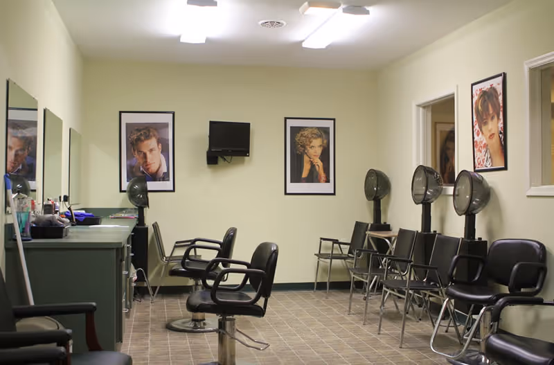 Interior view of a salon area with multiple black salon chairs and hair dryers. There are framed pictures of people on the walls and mirrors above a green counter with various hair styling tools. The floor is tiled and the room is well-lit with ceiling lights.