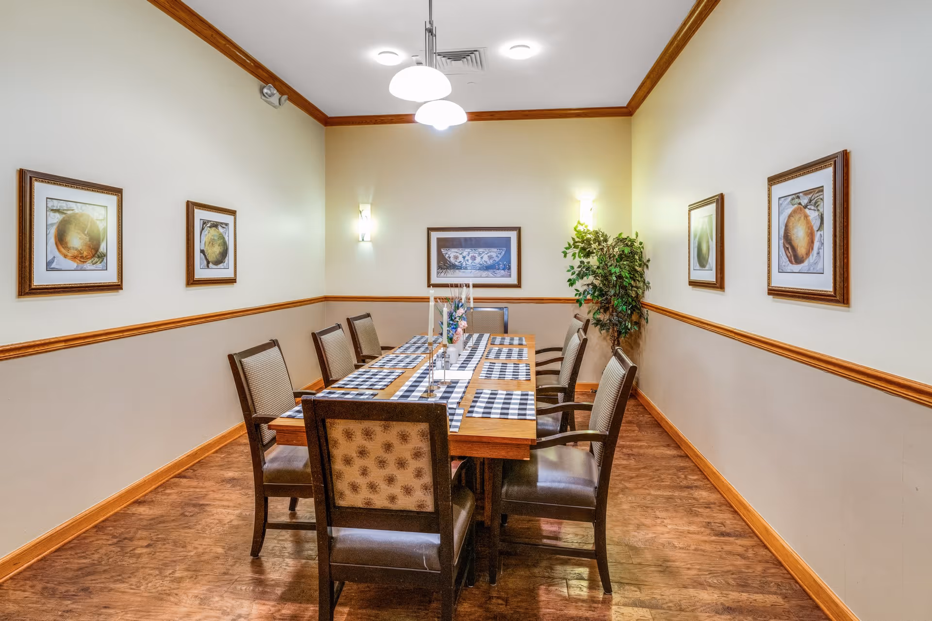 A dining room with a rectangular wooden table set for eight people with checkered placemats and a centerpiece. The room has wooden flooring, beige walls with wooden trim, framed artwork on the walls, and a potted plant in the corner. Ceiling lights illuminate the space.
