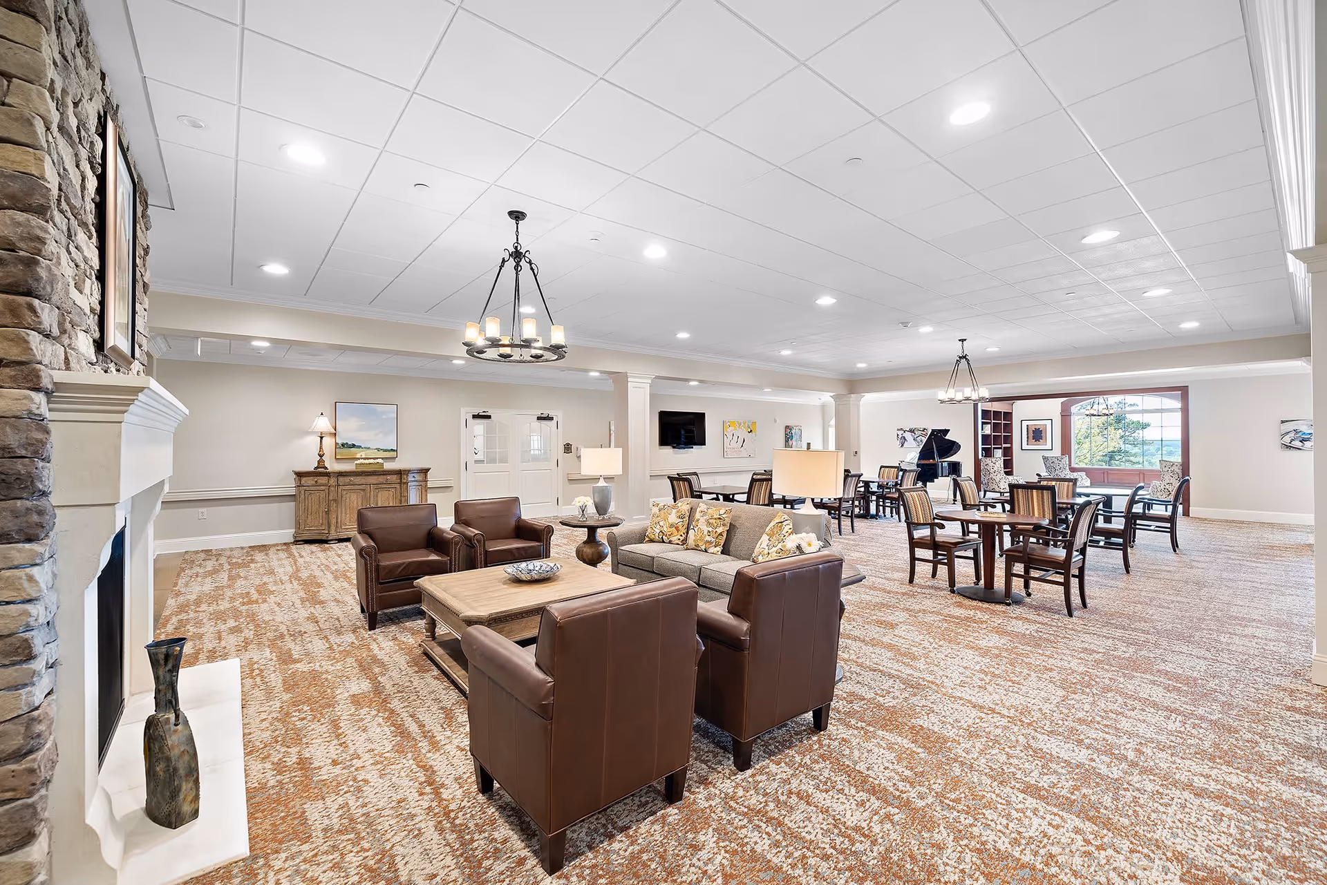 A spacious and well-lit common area in a senior living facility featuring a stone fireplace on the left, a seating area with a sofa and four leather armchairs around a wooden coffee table, multiple tables and chairs for dining or activities, a grand piano near a large window with a scenic view, and decorative lighting fixtures on the ceiling.