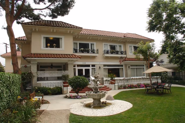 Exterior view of a two-story beige building with a red tile roof, surrounded by a well-maintained garden with green grass, flowers, bushes, and a multi-tiered stone fountain in the foreground. There is a patio area with a table, chairs, and an umbrella on the right side. Trees frame the scene on both sides.
