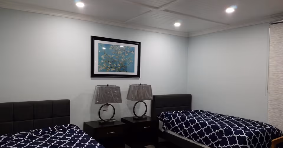 A bedroom with two single beds featuring dark upholstered headboards and blue and white patterned bedspreads. Between the beds are two dark wooden nightstands, each with a modern lamp with a circular base. Above the nightstands hangs a framed artwork depicting a branch with white flowers on a blue background. The room has light-colored walls, recessed ceiling lights, and a window with closed blinds on the right side.