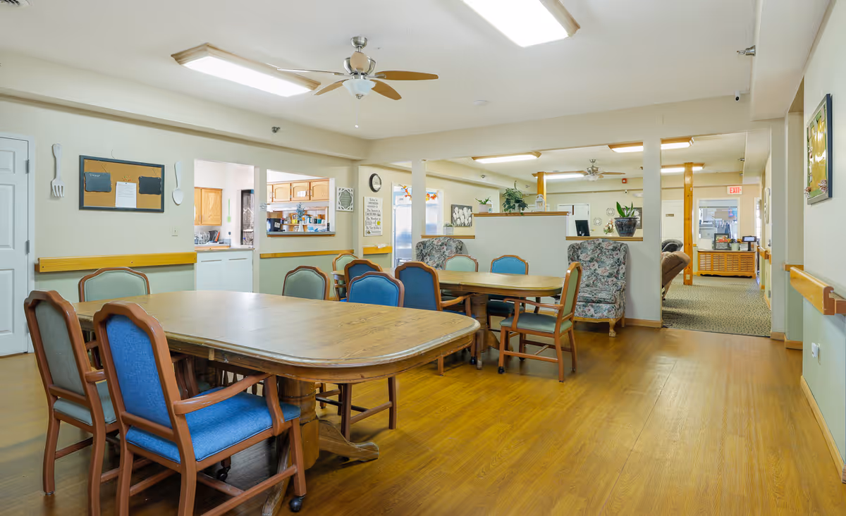 A spacious dining area in a senior living facility with wooden tables and chairs featuring blue and green upholstery. The room has light-colored walls, wood flooring, ceiling fans, and fluorescent lighting. There is a kitchen pass-through window on one side and a seating area with floral armchairs in the background. Various decorations and plants are visible, along with an exit sign in the far background.