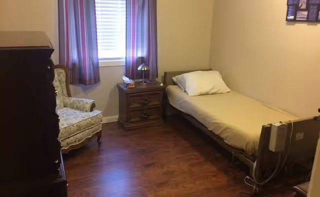A small bedroom with a single bed against the right wall, a wooden nightstand with a lamp and tissue box next to the bed, a floral upholstered armchair near the window with striped curtains, and a wooden dresser on the left side. The floor is wooden and the walls are light-colored.