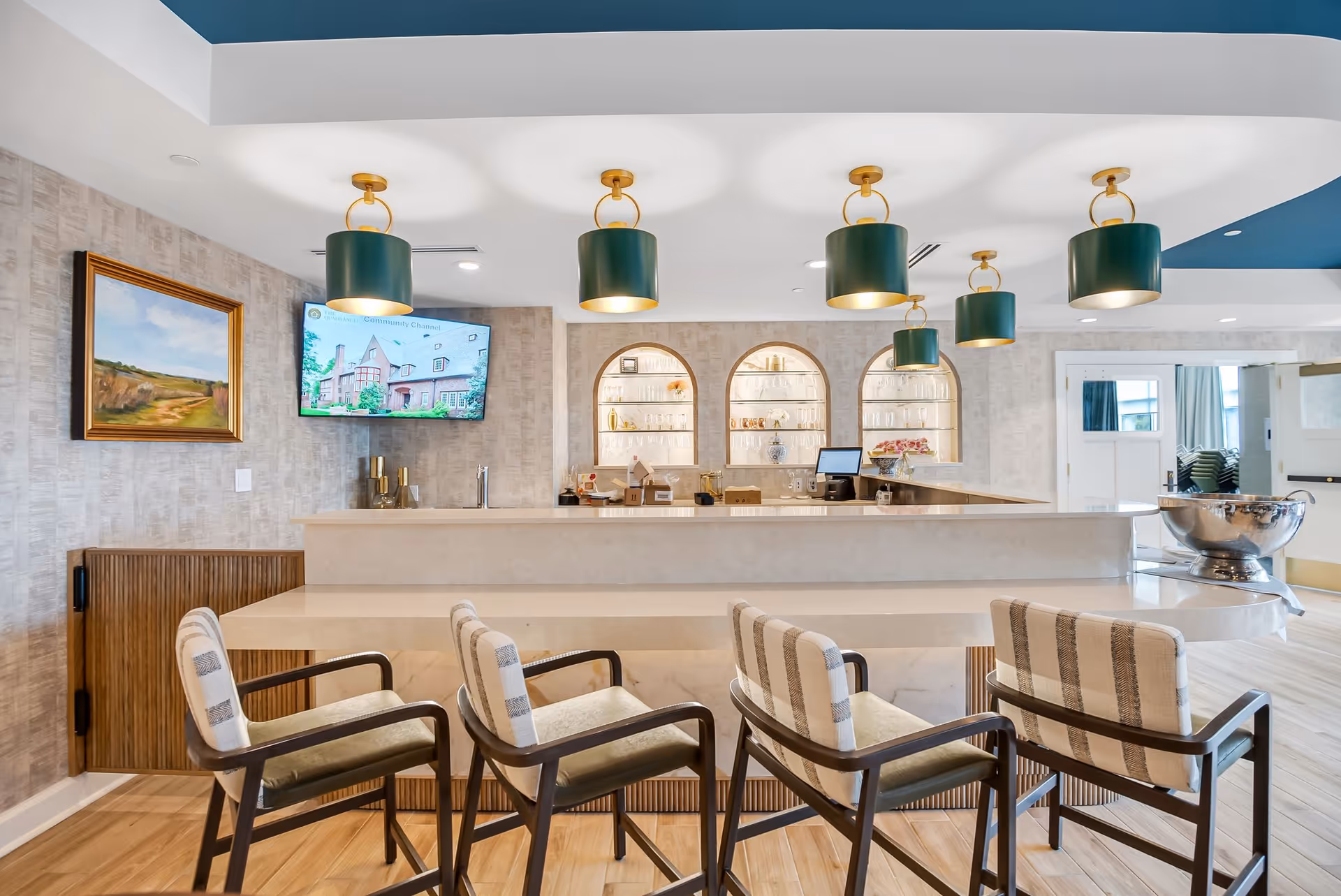 A modern and stylish bar area in a senior living facility with a marble countertop and four cushioned bar stools with wooden frames. Above the bar are five green and gold pendant lights. Behind the bar are three arched shelves with glassware and decorative items, a computer monitor, and a TV mounted on the wall displaying an image of a building. The walls have a textured beige wallpaper, and there is a framed landscape painting on the left wall. The floor is light wood, and the ceiling has a blue accent.