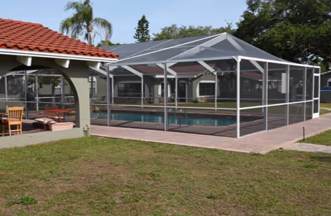 A screened-in outdoor swimming pool beside a stucco building with arched openings and a red tile roof, with grass in the foreground.