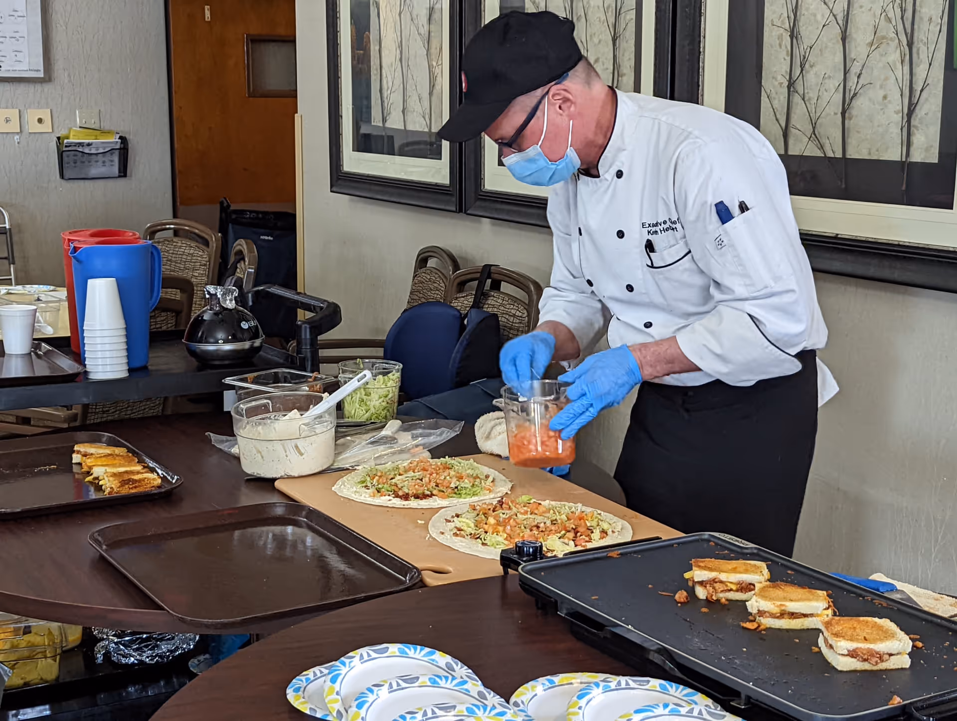 A chef wearing a white coat, black hat, blue gloves, and a face mask is preparing food on a table. There are two tortillas being topped with ingredients like lettuce and salsa. On the table, there are also containers with other ingredients, a tray with grilled sandwiches, disposable plates, and cups. The setting appears to be a dining or common area with chairs and framed pictures on the wall.