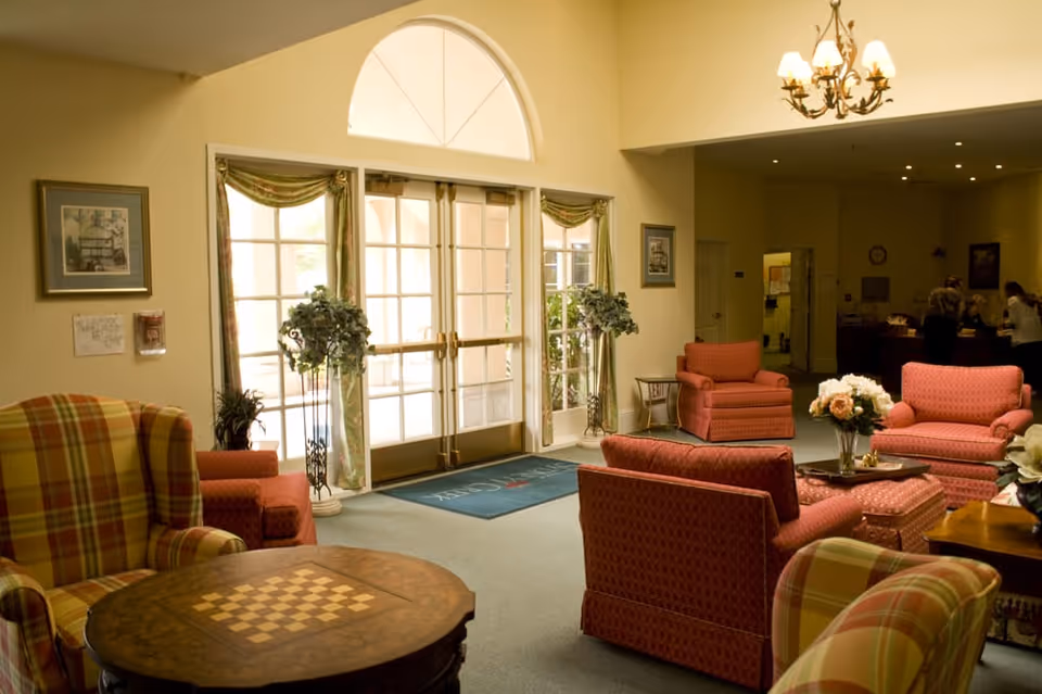 A cozy living room area with multiple upholstered armchairs and sofas in warm tones, a round wooden table with a chessboard pattern, floral arrangements on tables, large glass double doors with curtains letting in natural light, and a chandelier hanging from the ceiling. In the background, a reception desk with people standing behind it is visible.