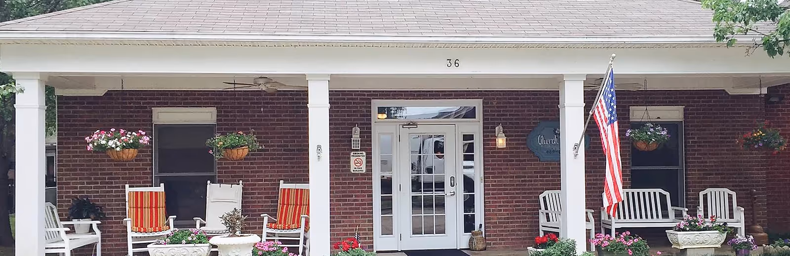 Front entrance of a brick assisted living building with a covered porch, white columns, seating, hanging flower baskets, and an American flag.