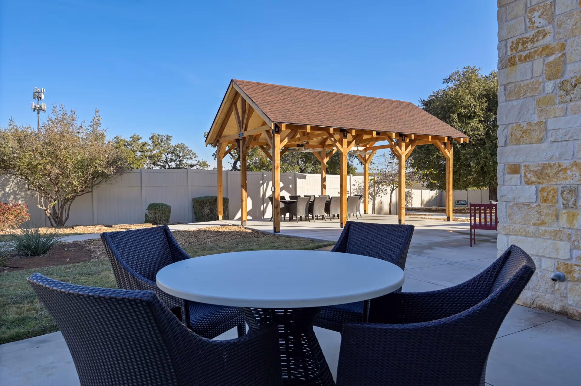 Outdoor seating area with a round table and four wicker chairs in the foreground, and a wooden pavilion with a pitched roof and additional seating in the background, surrounded by trees and a white fence under a clear blue sky.