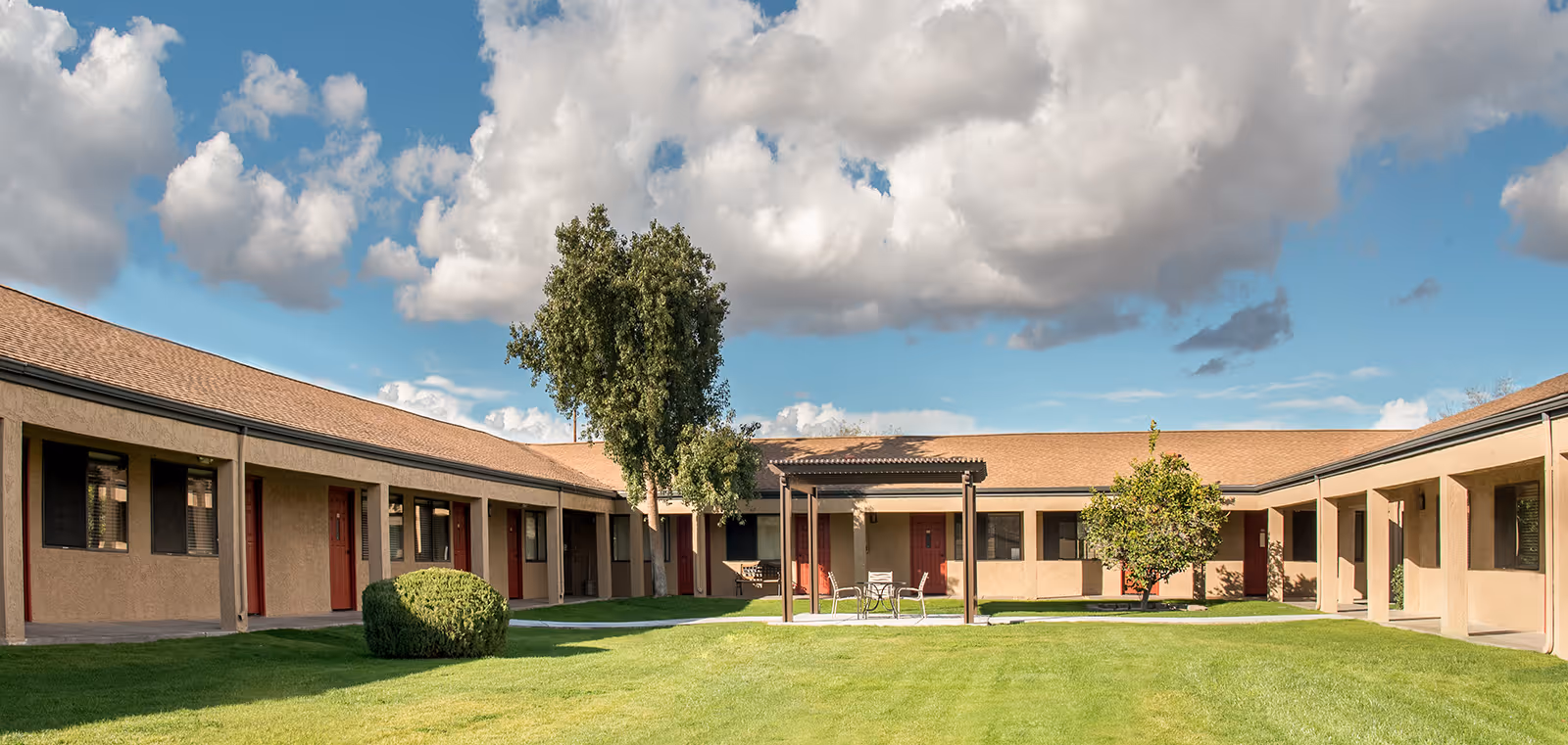 A courtyard area of Paseo Village senior living facility featuring a green lawn, a few trees, a small pergola with a table and chairs underneath, and a single-story building with multiple red doors and windows surrounding the courtyard.