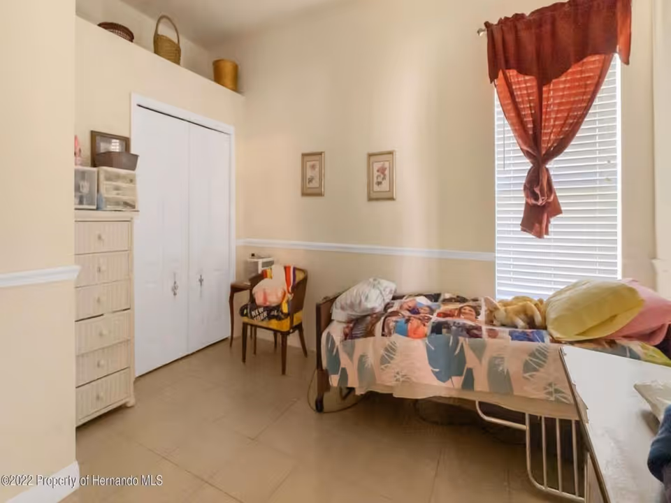 A small bedroom with a single bed covered with a colorful blanket and pillows. There is a window with closed blinds and a red curtain tied in the middle. A wooden chair with a blanket draped over it is next to the bed. A white dresser with multiple drawers is on the left side near a closed white closet door. The walls are painted light beige with a white chair rail molding.