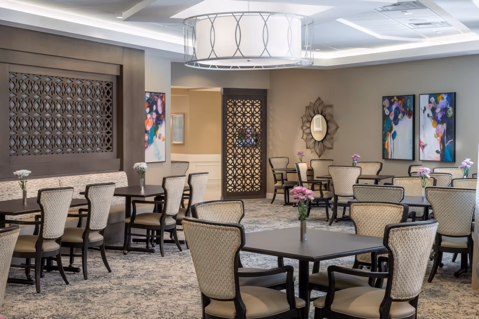 A spacious dining room with multiple tables and cushioned chairs arranged neatly. Each table has a small vase with flowers. The room features decorative wall panels, colorful abstract paintings, a decorative mirror, and a large modern ceiling light fixture. The carpet has a patterned design, and the walls are painted in neutral tones.