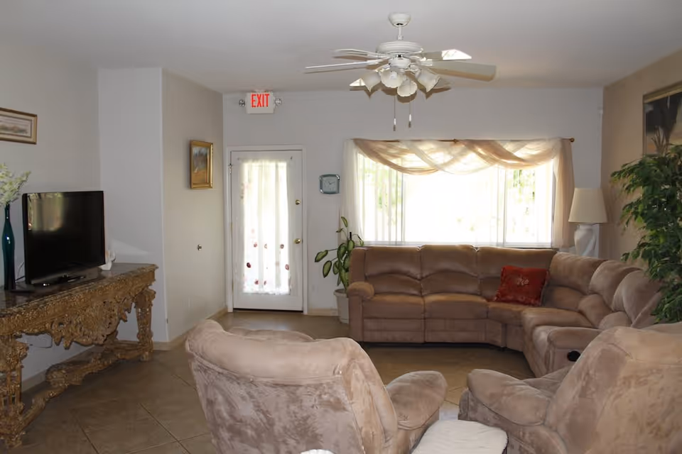 A cozy living room with a large beige sectional sofa and two matching armchairs arranged around a TV on an ornate wooden console table. There is a ceiling fan with lights, a window with sheer curtains, a potted plant, a floor lamp, and an exit door with a sheer curtain.
