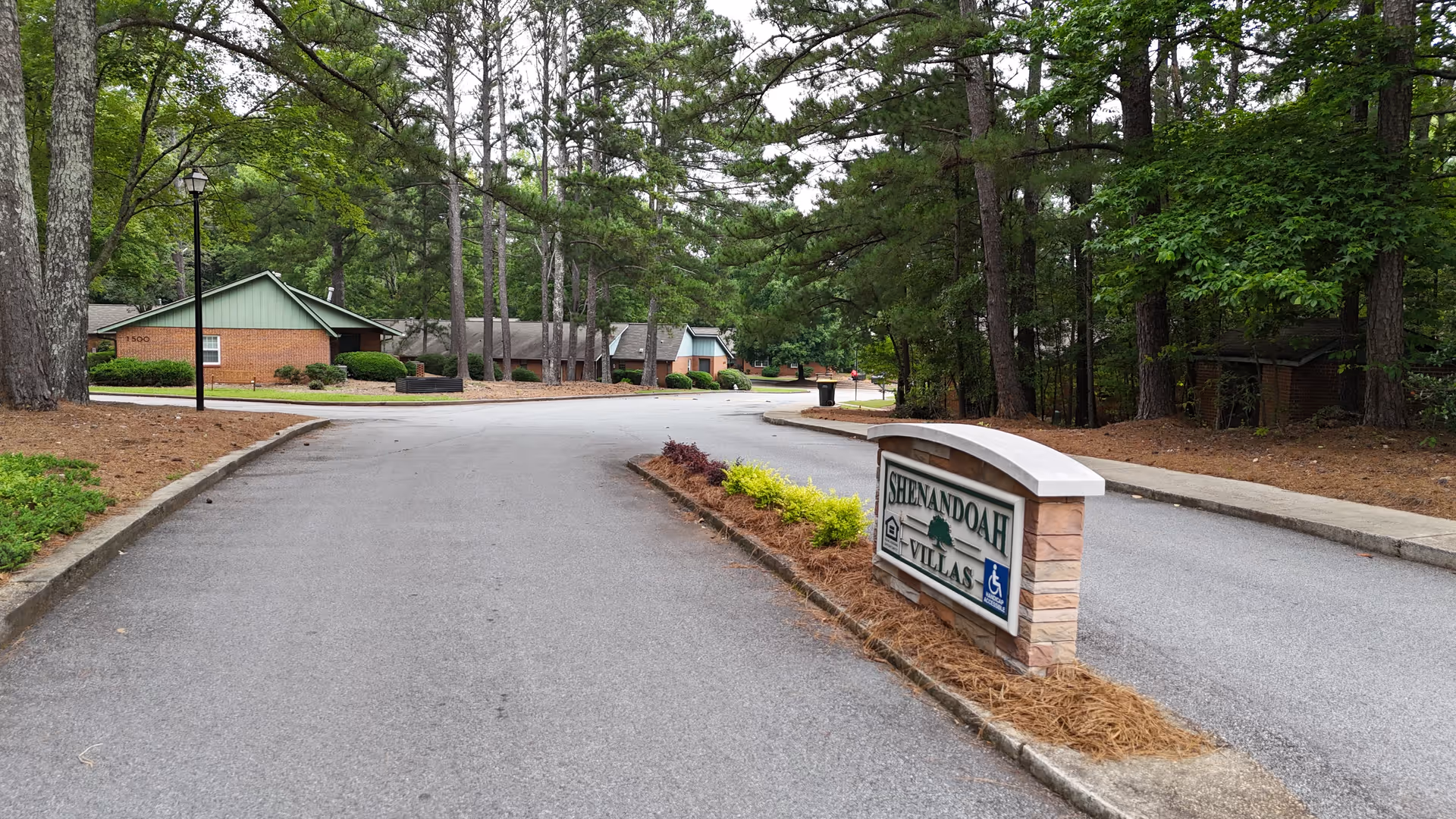 Entrance road to Shenandoah Villas senior living facility with a stone sign displaying the facility name. The road is lined with trees and shrubs, and single-story brick buildings with green roofs are visible in the background.