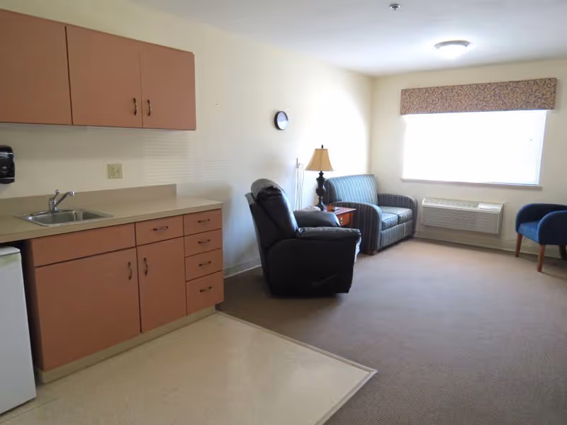 A small living area with a kitchenette on the left featuring cabinets, a sink, and a mini refrigerator. The living space includes a black recliner, a striped loveseat, a wooden side table with a lamp, a blue chair, a wall clock, and a window with a patterned valance. The floor transitions from linoleum in the kitchenette to carpet in the living area.