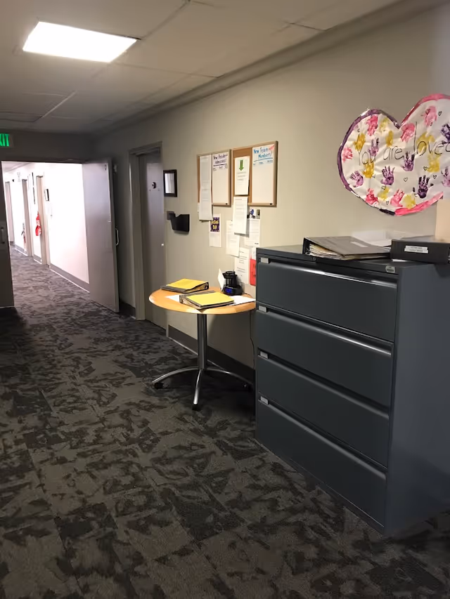 Interior hallway with patterned carpet, a grey filing cabinet and small round table holding folders beneath a bulletin board and heart-shaped artwork.