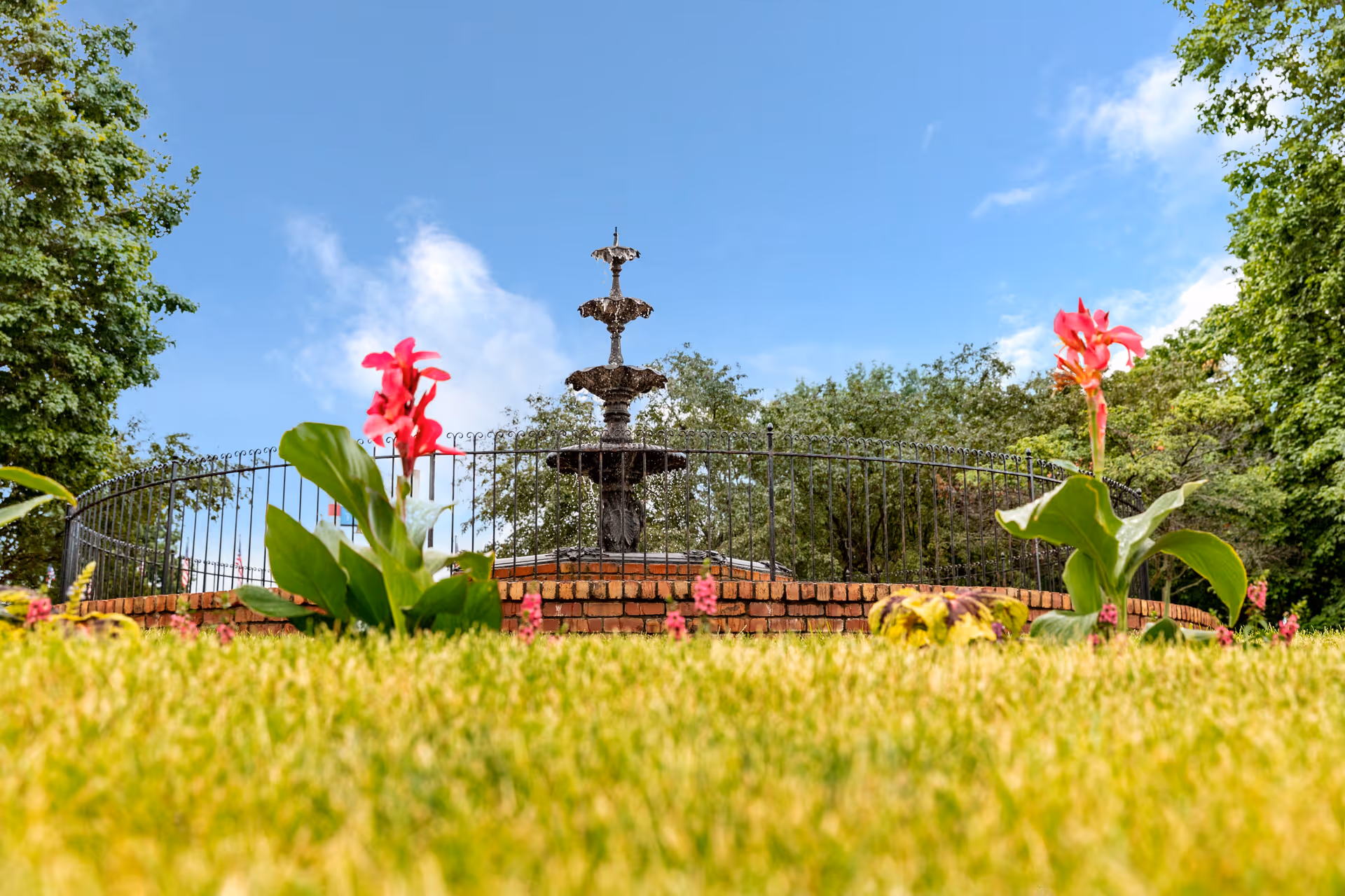 Low-angle view of a garden with green grass and red flowers in the foreground, a black wrought iron fence, and a multi-tiered water fountain surrounded by trees under a blue sky with some clouds.