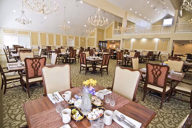 Spacious dining room with multiple tables and chairs arranged neatly. Tables are set with placemats, cups, glasses, utensils, and condiments. The room features high ceilings with chandeliers and a balcony area overlooking the dining space. Large windows allow natural light to brighten the room.