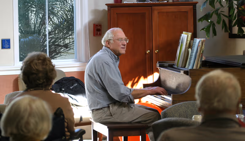 An elderly man playing a piano in a well-lit room with several elderly people seated and watching him. The room has a large window with a view of greenery outside, a wooden cabinet, and some plants.